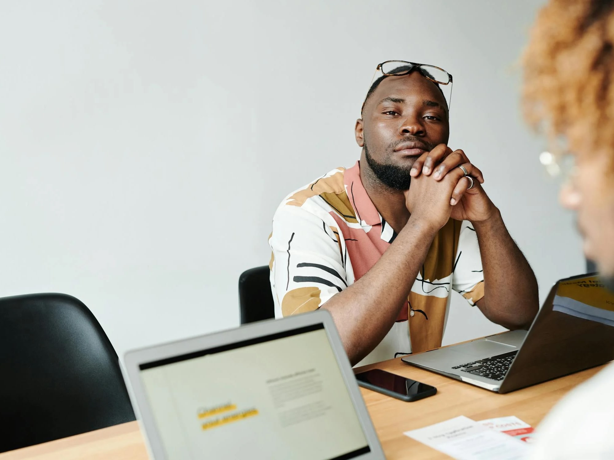 Man in patterned shirt with glasses on forehead sitting at table with laptop and phone, facing another person.