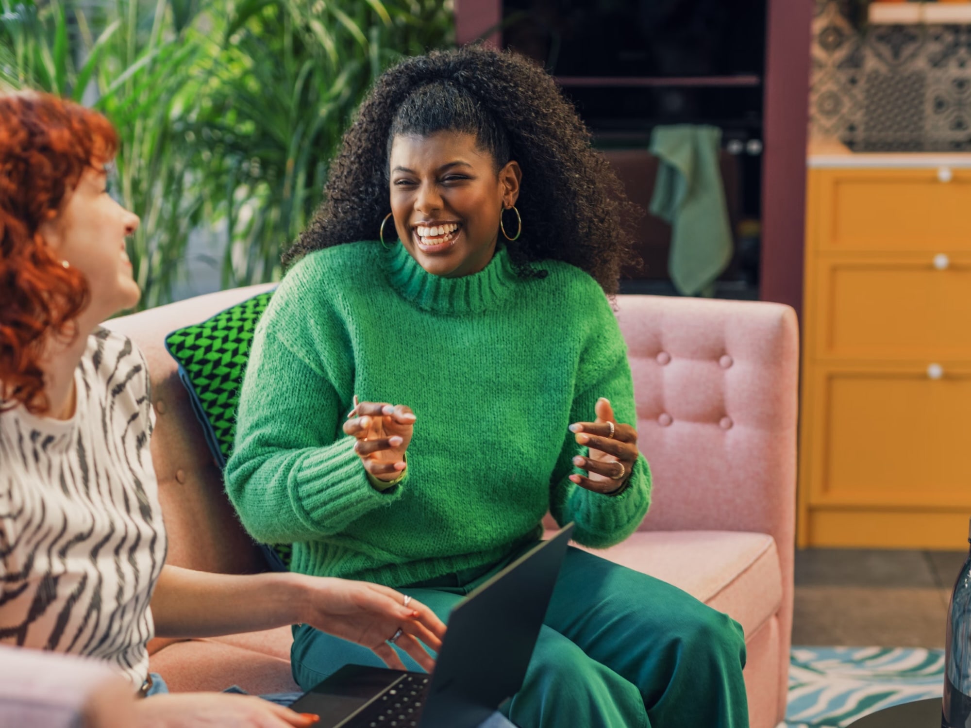 Two women chatting and smiling on a pink couch, one wearing a green sweater and holding a laptop.
