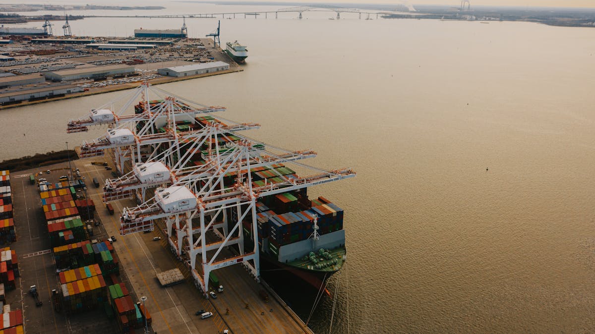 Aerial view of a cargo ship docked at a port with large cranes and stacked shipping containers.