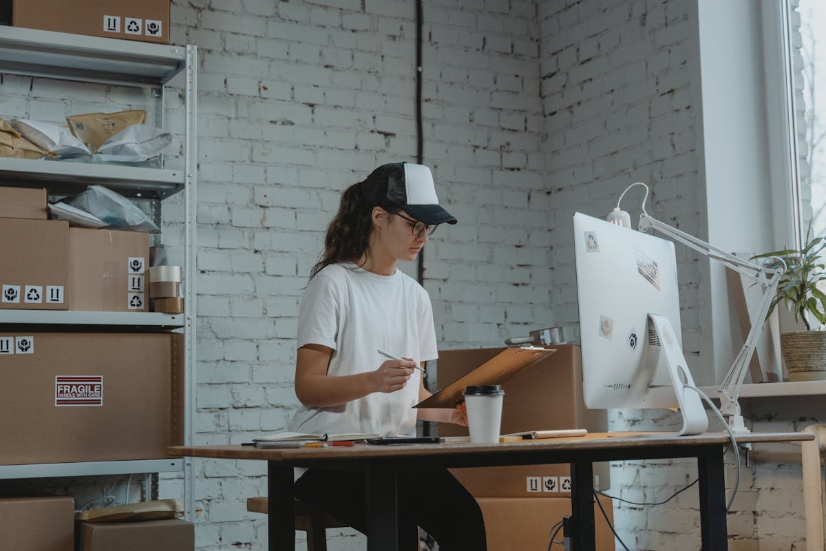 Woman in cap and glasses working on a clipboard at a desk with computer and packages nearby, warehouse setting.