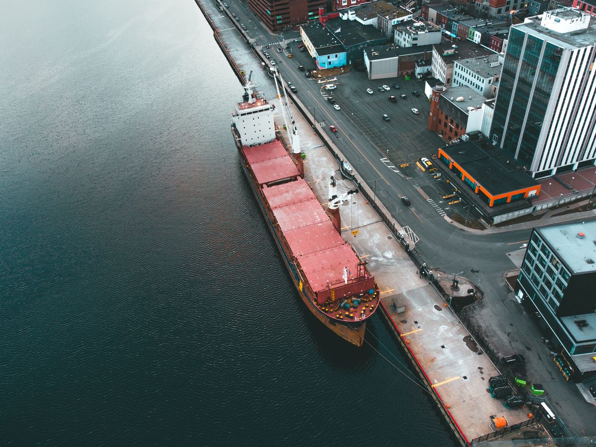 A large cargo ship docked at a port beside an urban area with buildings and parked cars.