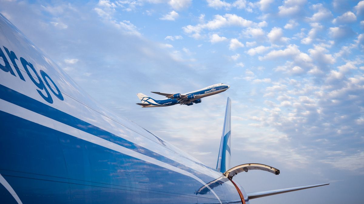 Cargo plane wing with open hatch and another cargo plane taking off against cloudy blue sky.