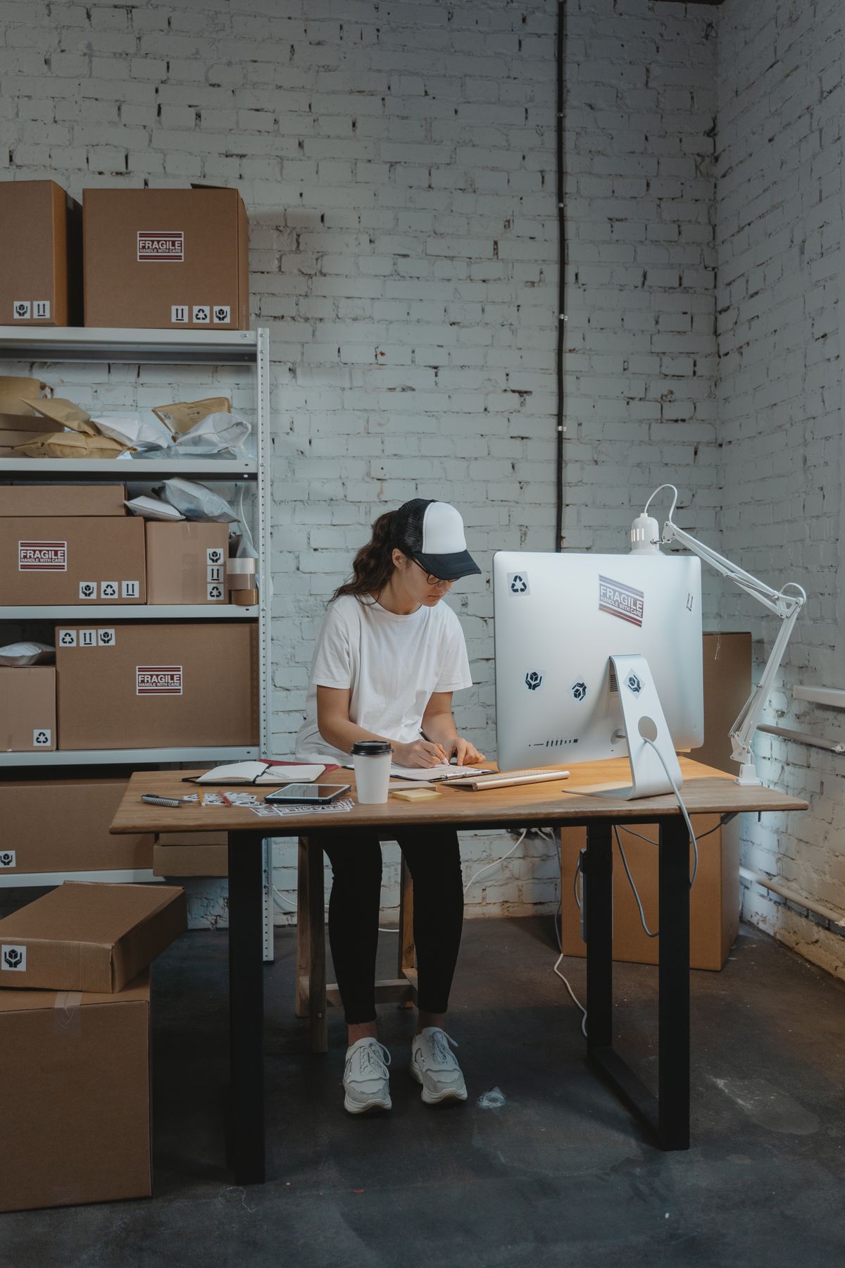 Woman in cap writing at a desk with computer and coffee, surrounded by cardboard boxes in warehouse.