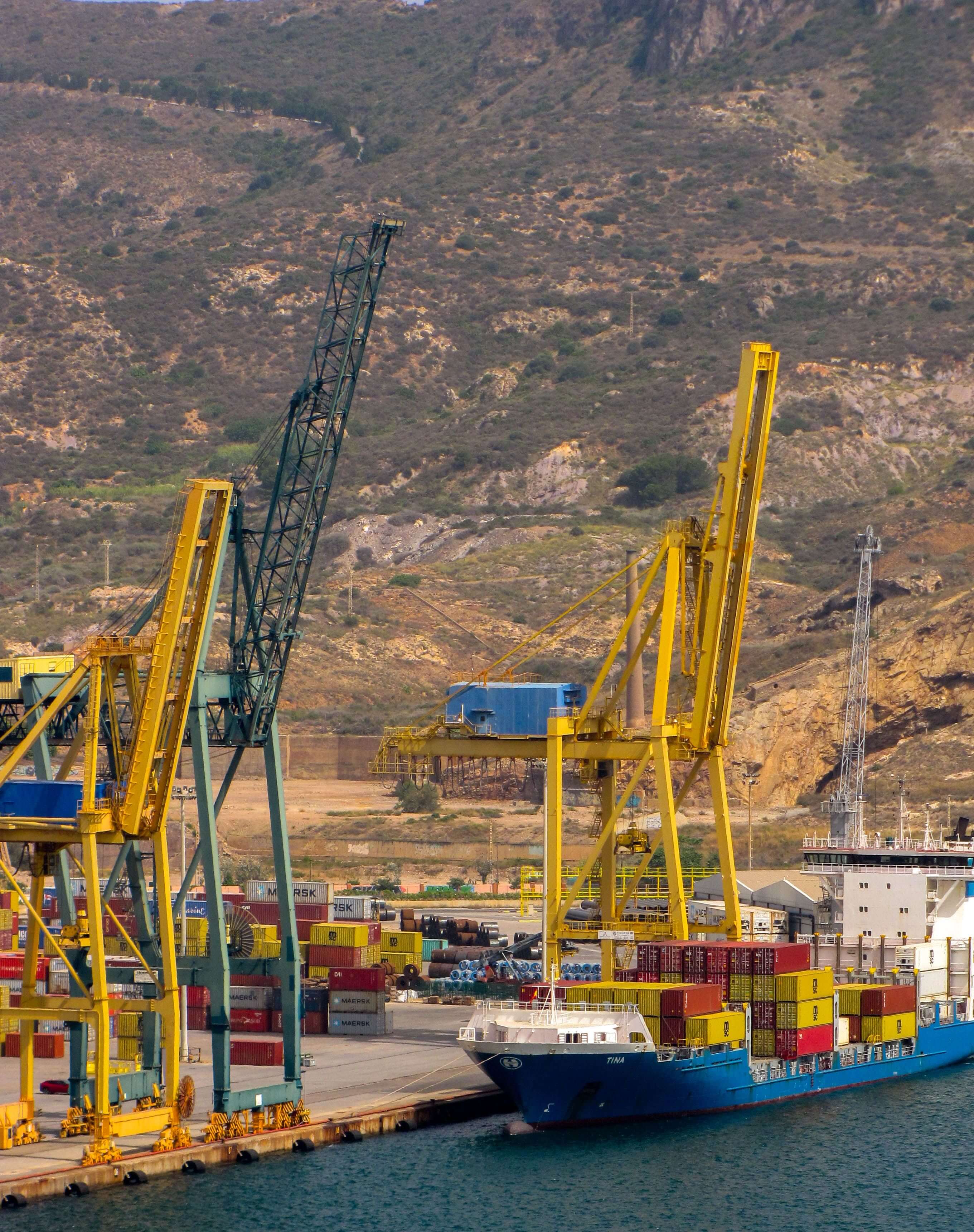 Cargo ship named Tina docked at port with yellow cranes and stacked shipping containers.