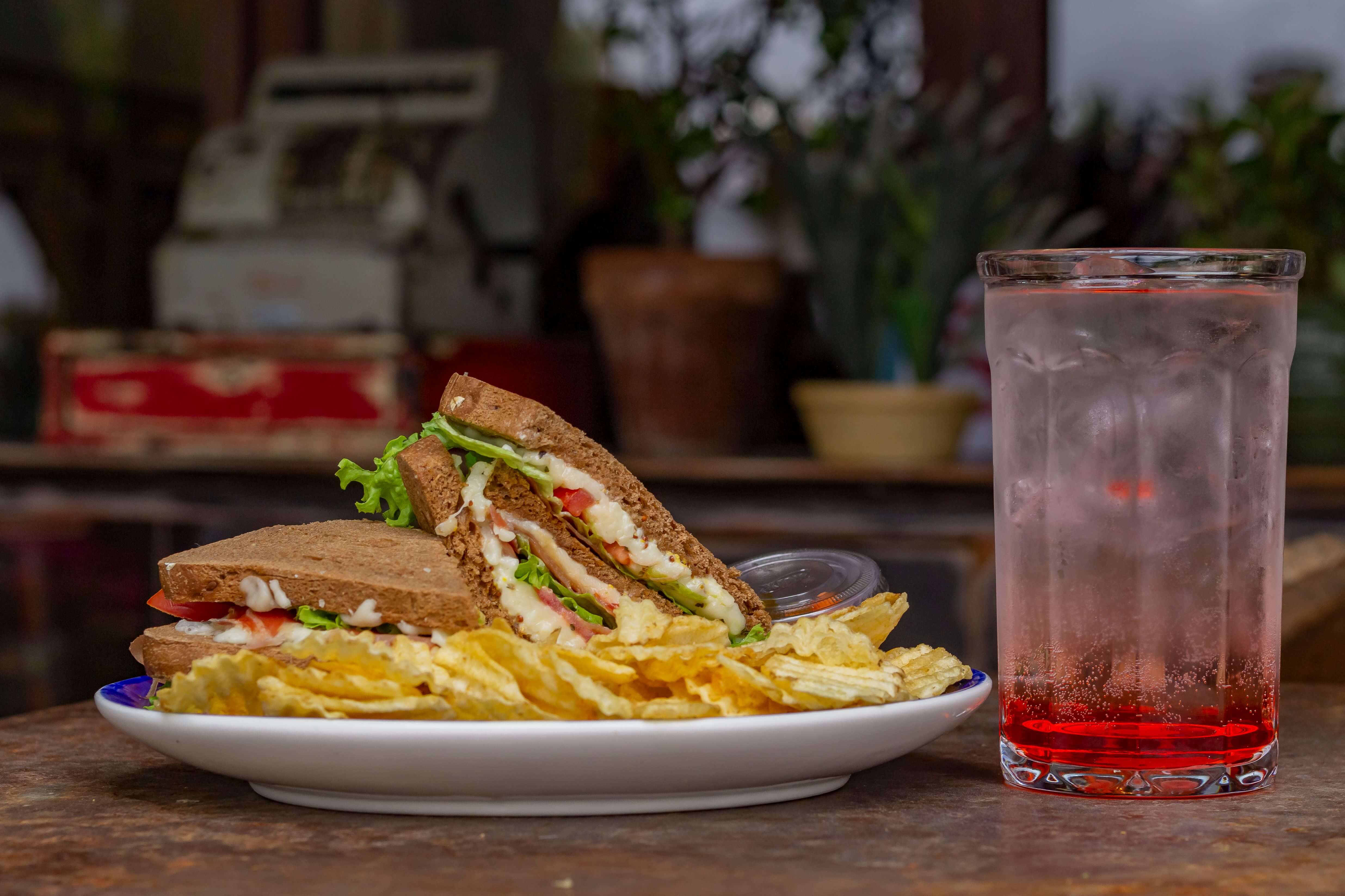 Plate with sandwich, potato chips, and a glass of red soda with ice on a rustic table.