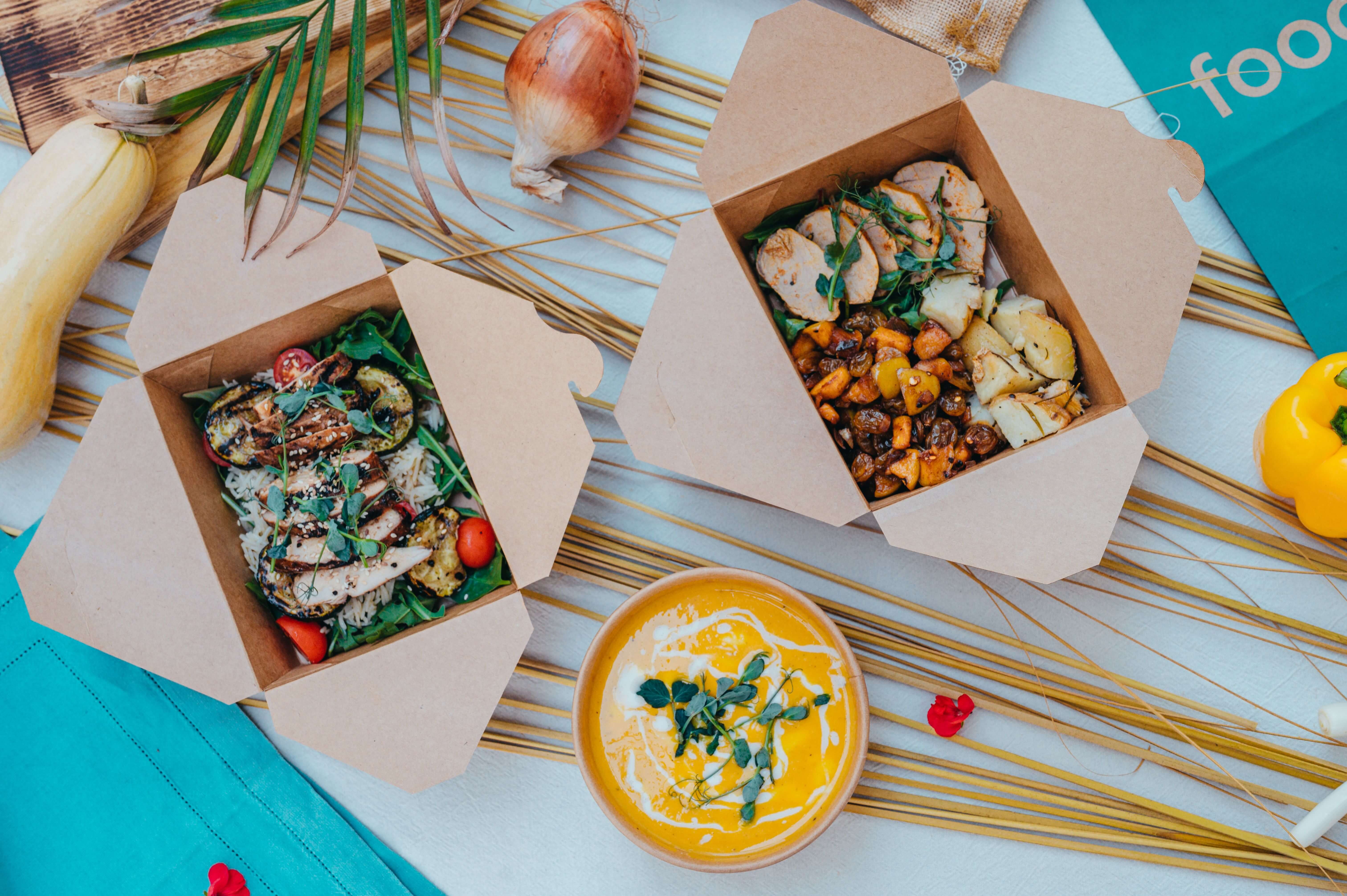 Two cardboard takeout boxes with grilled chicken salads and a bowl of creamy soup garnished with herbs.