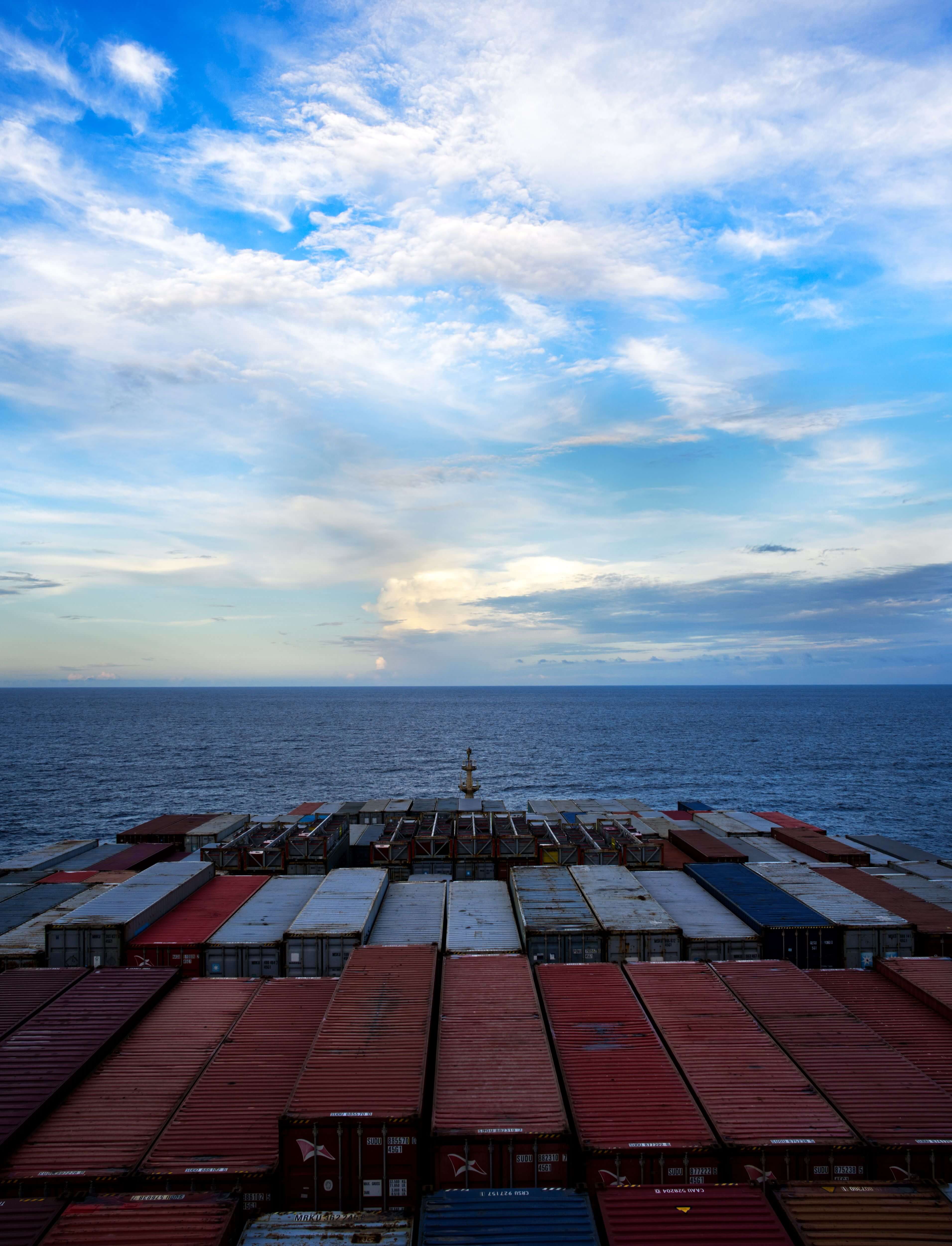 View over stacked shipping containers on a cargo ship with ocean and partly cloudy sky.