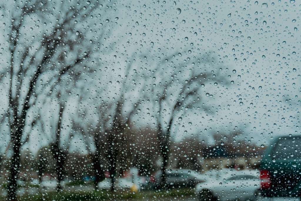 Raindrops on a window with blurred trees and cars outside.