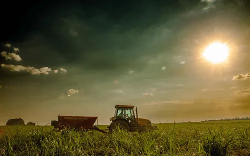 Tractor pulling an implement on a green field under a sunny sky with scattered clouds.