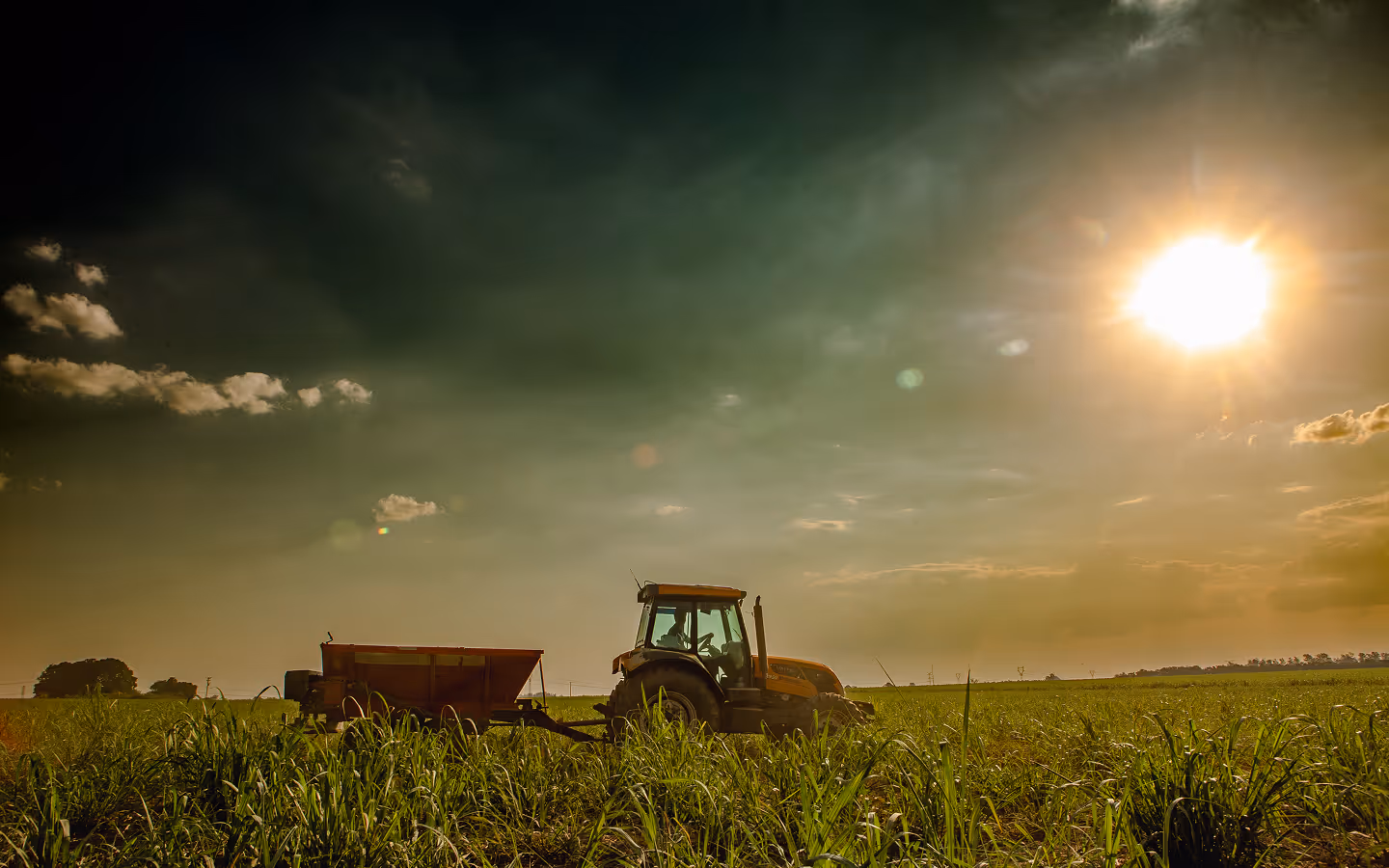Tractor pulling an implement on a green field under a sunny sky with scattered clouds.