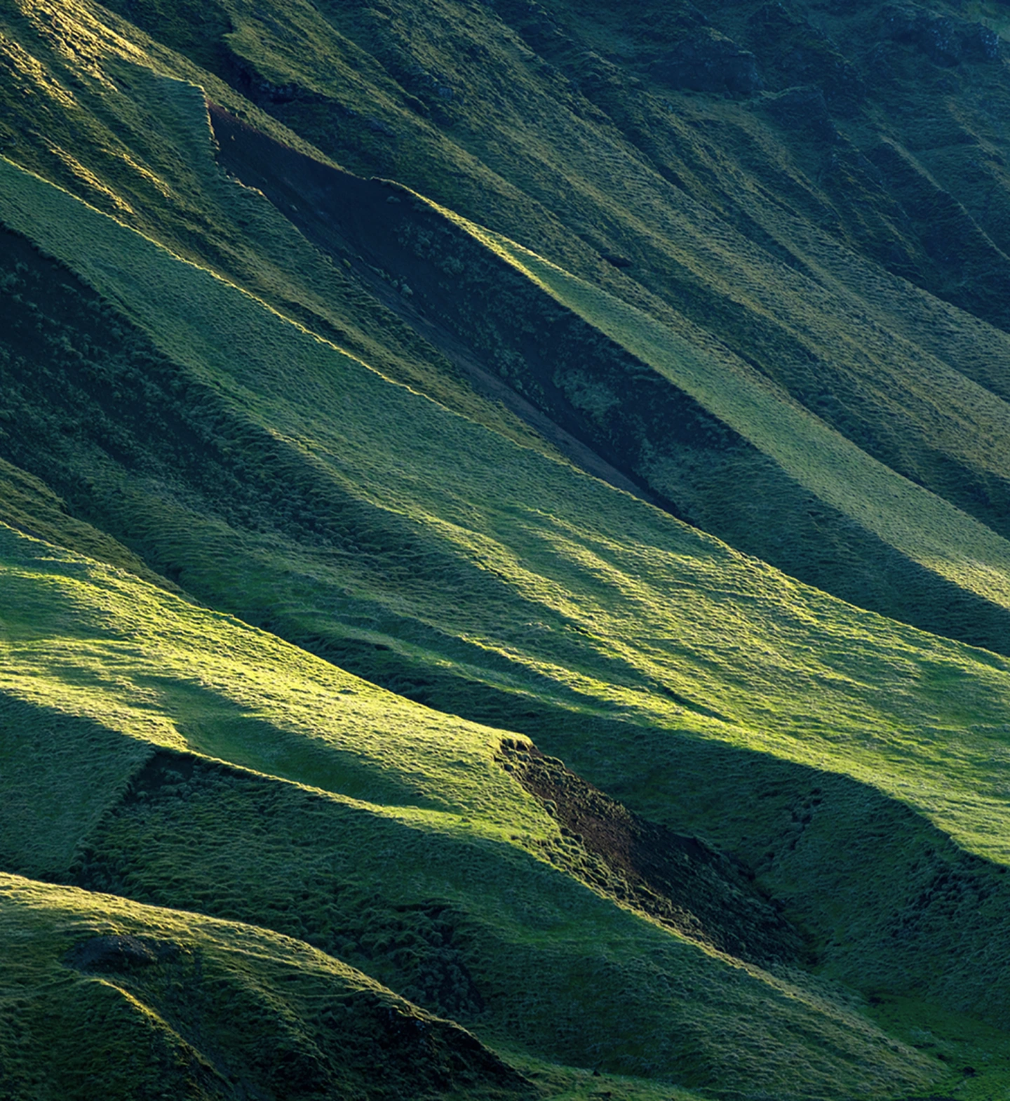Sunlight casts shadows on lush green rolling hills with steep slopes and ridges.