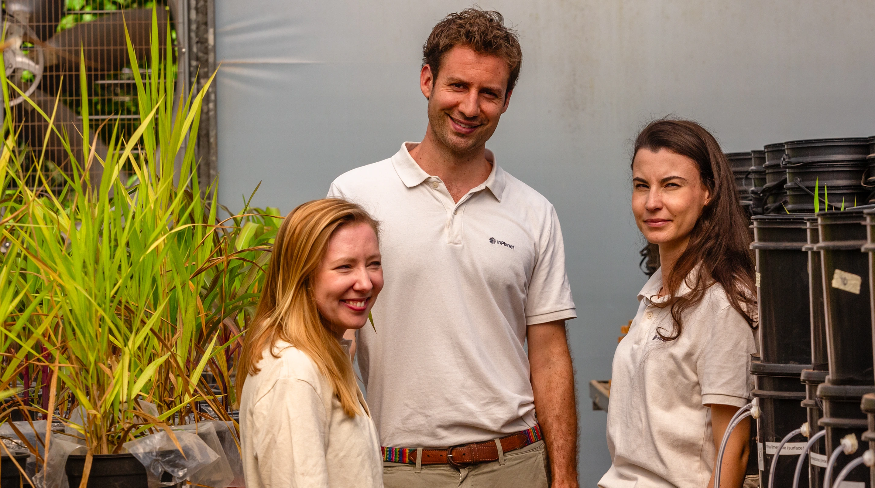 Three smiling people in white shirts stand among plants and stacked black containers indoors.