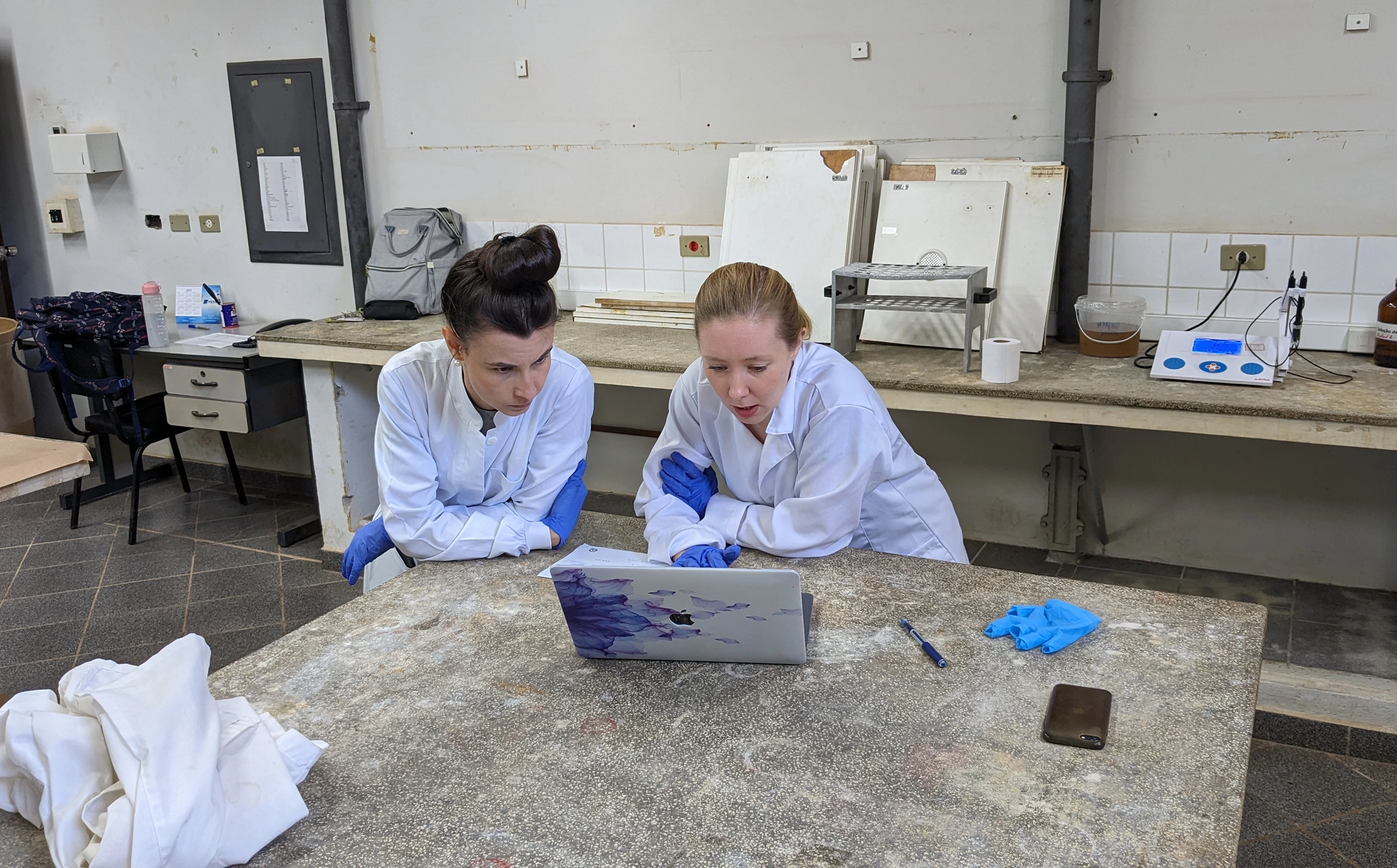 Two female scientists in white coats and gloves studying a laptop in a lab with equipment around them.