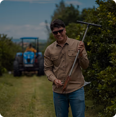 Man with sunglasses holding a farming tool standing on a field with a blue tractor in the background.