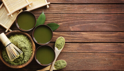 Aufsicht auf auf Holzdielen mit frischen Teeblättern und Teezeremonie-Utensilien : Drei japanische Teeschalen gefüllt mit grünem Tee, Schale und Holzlöffel mit Matcha-Pulver, Matcha-Besen