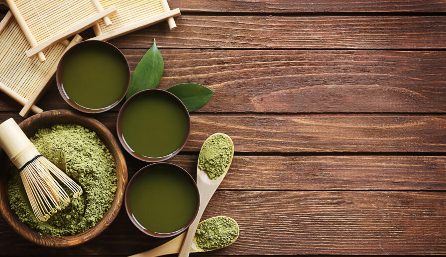 Aufsicht auf auf Holzdielen mit frischen Teeblättern und Teezeremonie-Utensilien : Drei japanische Teeschalen gefüllt mit grünem Tee, Schale und Holzlöffel mit Matcha-Pulver, Matcha-Besen