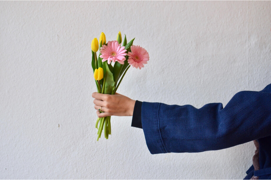 Weiblicher Arm in blauer Jacke, der von rechts ins Foto ragt, so als werde er jemandem entgegengestreckt.Die Hand hält einen bunten Blumenstrauß