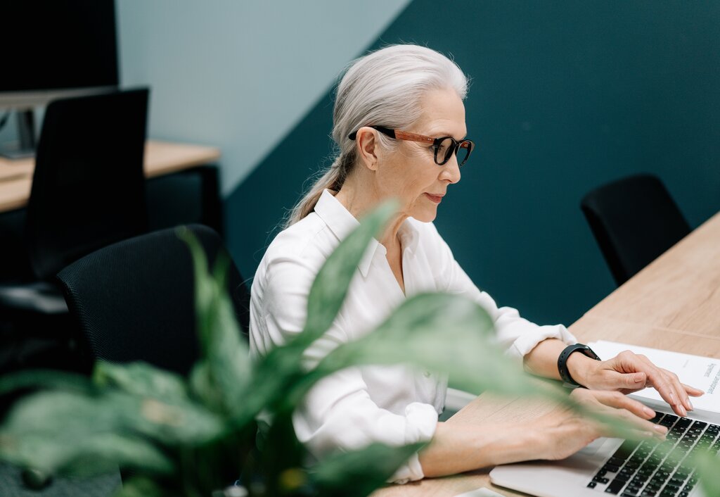 Sportlich-elegante ältere Dame mit vollem zu einem Nackenzopf gebundenen weißem Haar, schlichter weißer Bluse und Brille am Schreibtisch im Büro auf der Computer-Tastatur schreibend