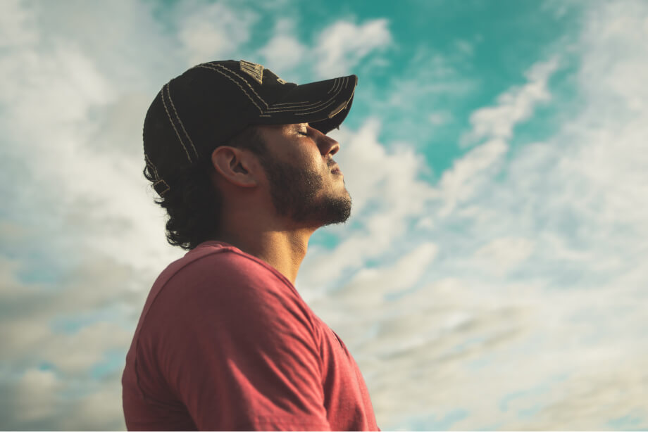 Junger Mann im Profil mit Baseball-Cap, kurzem Bart und T-Shirt vor blauem Himmel mit weißen Wolken. Kopf leicht in den Nacken gelegt, Augen genießerisch geschlossen, Sonnenstrahlen auf dem Gesicht
