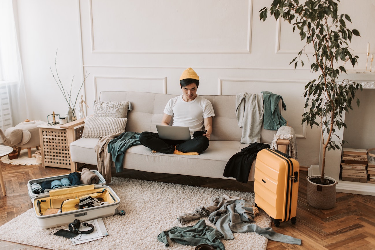 Ein junger Mann sitzt entspannt auf der Couch vor seinem ausgepackten Koffer und arbeitet am Laptop