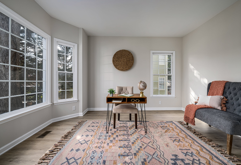 Gemütliches und modernes Wohnzimmer mit viel natürlichem Licht durch große Erkerfenster. Minimalistisch eingerichtet mit einem gewebten Wandbehang, einem stilvollen Schreibtisch mit Dekorationsgegenständen, einem grauen Plüschsofa mit rosa Überwurf und einem lebhaft gemusterten Teppich auf Holzboden.