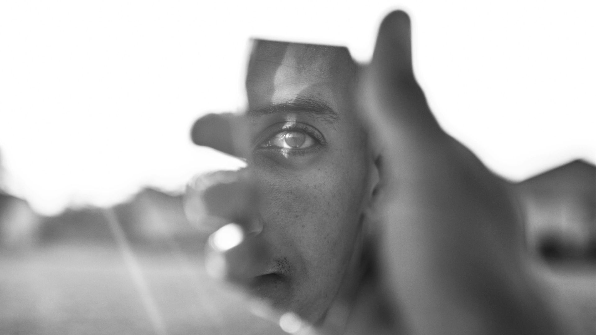 Nahaufnahme in Schwarz-Weiß eines jungen Mannes, der mit den Fingern sein Auge rahmt, mit Fokus auf die Spiegelung im Auge und unscharfem Hintergrund.