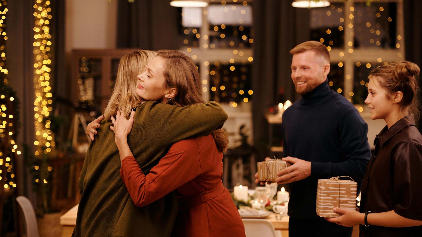 Gemütliches Weihnachtstreffen mit Freunden und Familie in festlicher Atmosphäre. Zwei Frauen umarmen sich herzlich, während im Hintergrund zwei Personen mit Geschenken lächeln. Warme Lichterketten und eine einladende Tischdekoration schaffen eine stimmungsvolle Weihnachtskulisse.