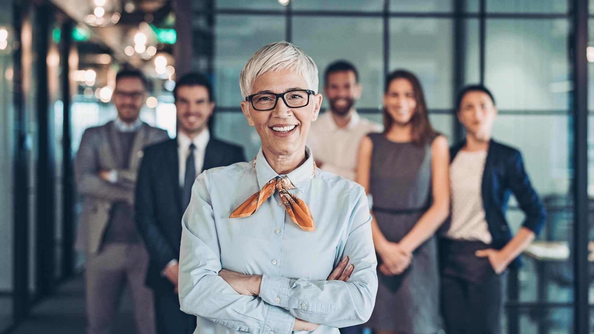 Eine selbstbewusste Führungskraft mittleren Alters mit weißem Haar und Brille steht lächelnd im Vordergrund, unterstützt von einem professionellen Team im Hintergrund. Perfekt für Themen wie Leadership, Teamarbeit, Unternehmenskultur und gesundes Arbeiten im Büro.