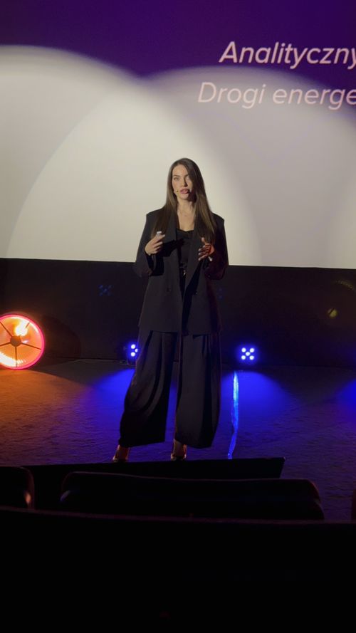 Kobieta na scenie mówiąca podczas prezentacji z oświetleniem i fragmentem napisu na ekranie.