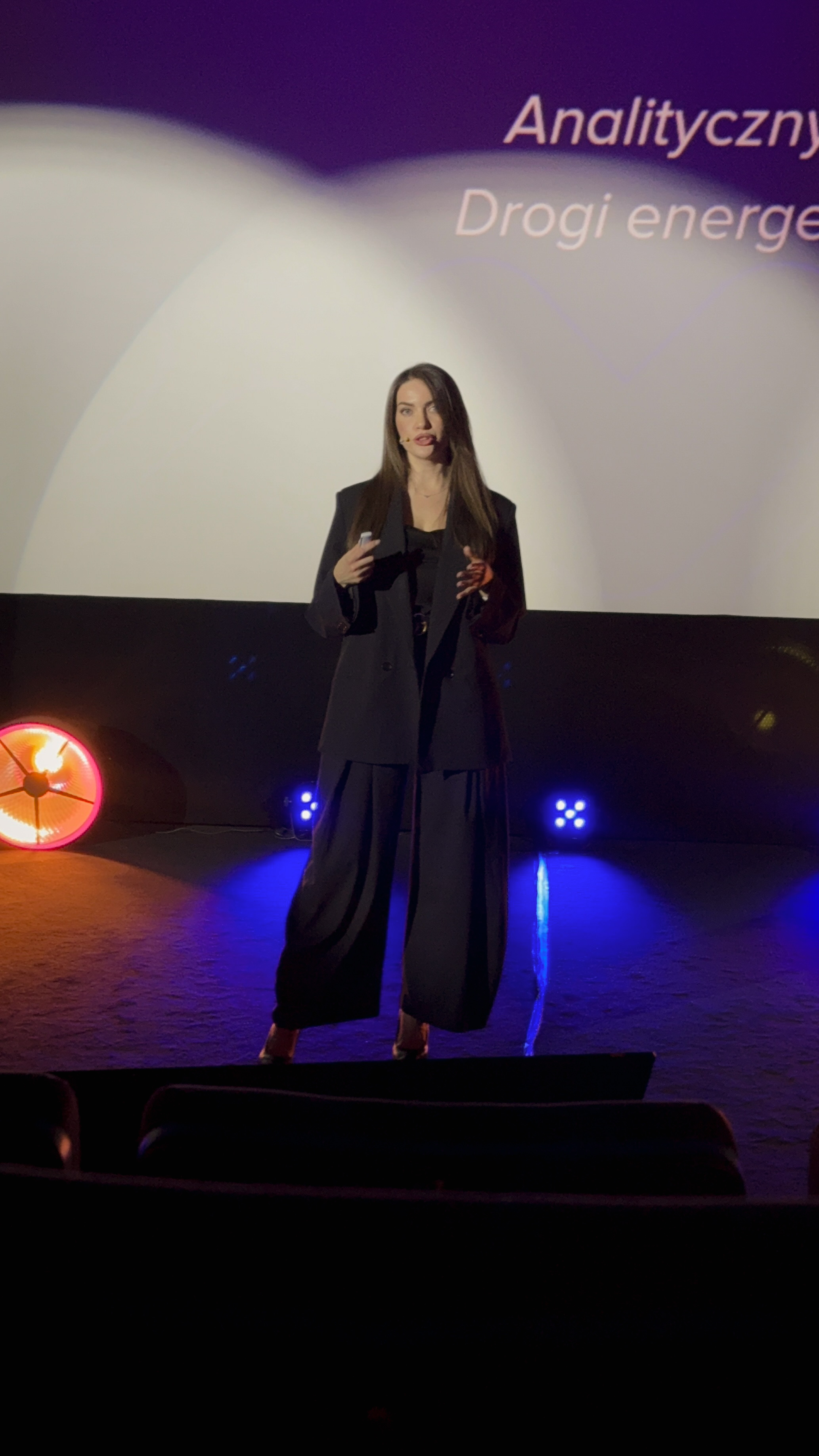 Kobieta na scenie mówiąca podczas prezentacji z oświetleniem i fragmentem napisu na ekranie.