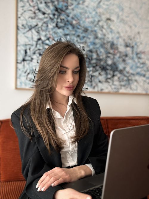 Kobieta w czarnej marynarce i białej koszuli pracująca na laptopie na pomarańczowej kanapie.