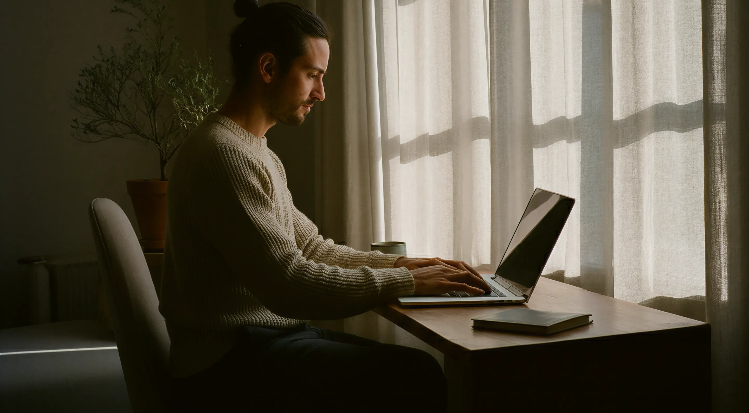 Man in sweater typing on a laptop at a wooden desk near a window with sheer curtains.