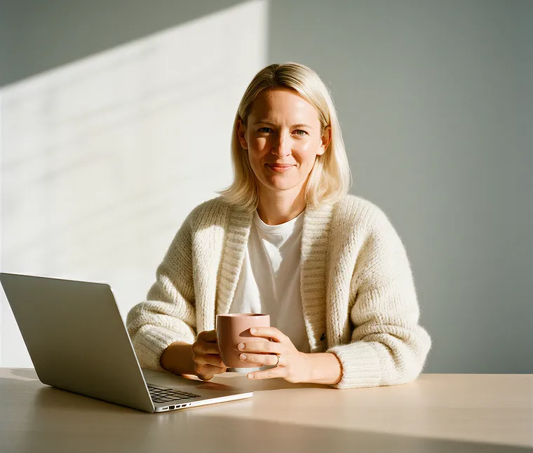 Woman with blonde hair sitting at table with laptop, holding a pink mug and smiling.