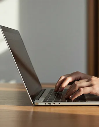 Person typing on a laptop keyboard on a wooden table in soft natural light.