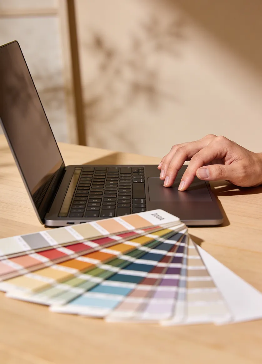 Hand using laptop touchpad near a color swatch fan on a wooden desk.