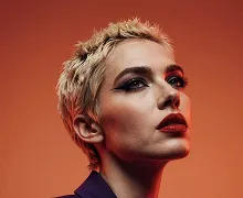Portrait of a person with short blond hair and bold makeup against an orange background.