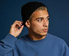 Man in blue sweatshirt and black knit hat touching the hat, looking to the side against a dark background.