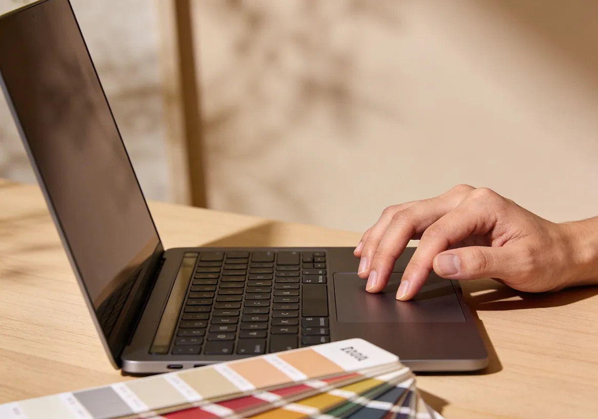 Person using a laptop touchpad on a wooden desk with a color swatch fan in the foreground.