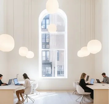 Four people working on laptops in a bright, modern office with large window and hanging globe lights.