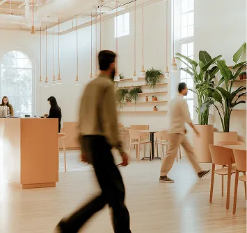 Two people walking inside a modern, spacious cafe with light wood furniture and indoor plants.