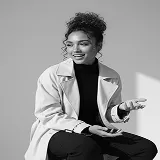 Smiling woman with curly hair in a light trench coat gesturing with hands against a plain background.
