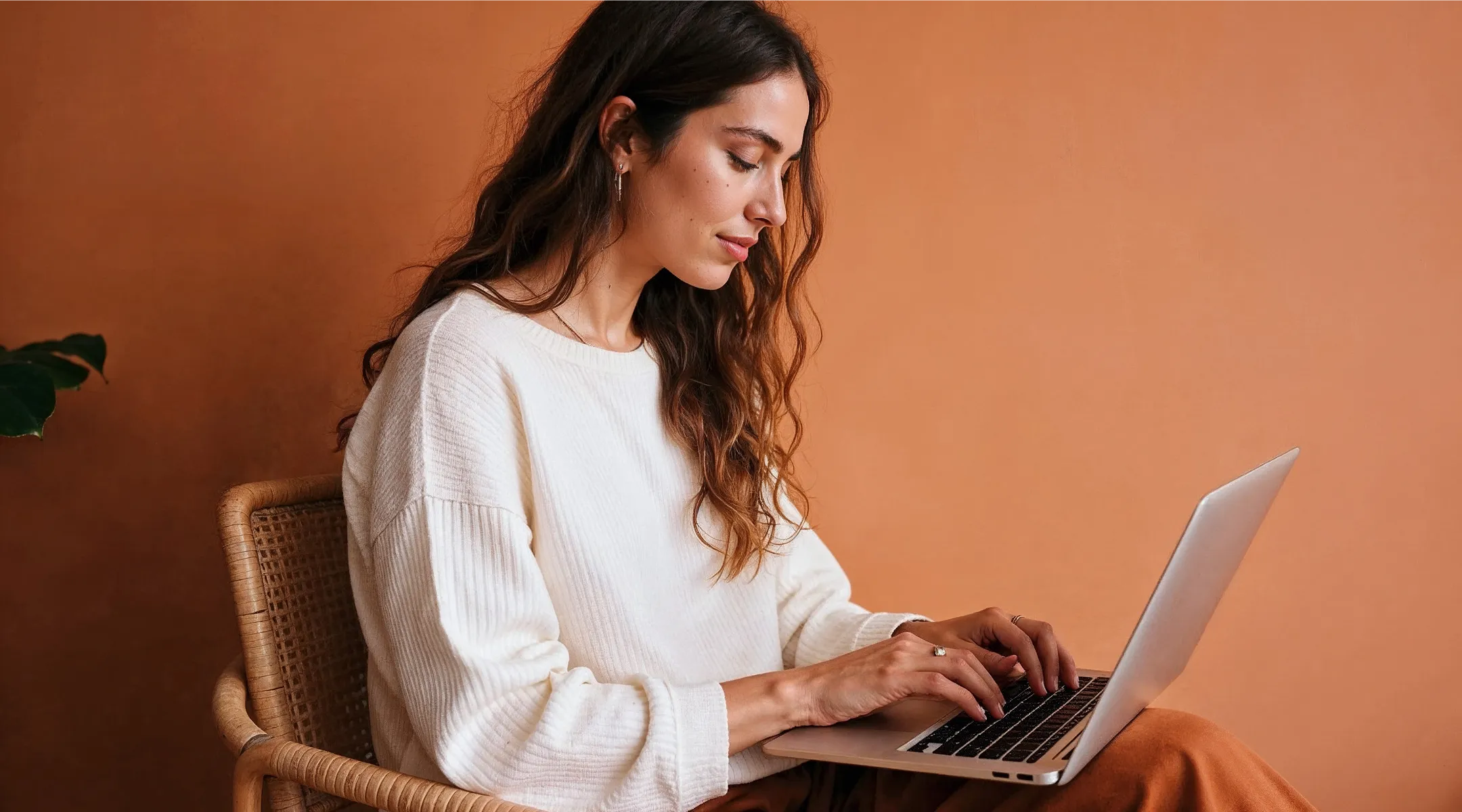 Woman with long wavy hair wearing white sweater typing on a laptop sitting on a wicker chair.