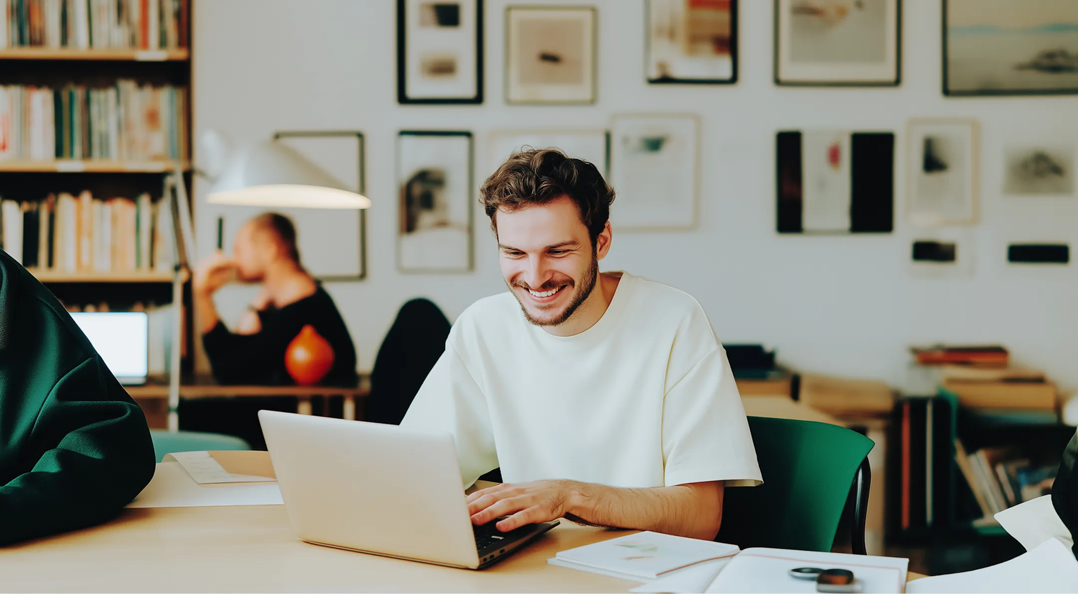 Smiling man in white shirt typing on laptop at desk in a cozy office with books and framed art.