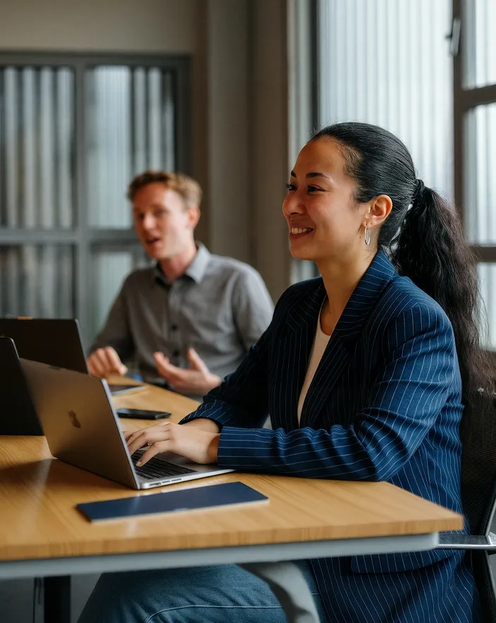 Smiling woman in pinstripe blazer typing on laptop at desk in office with male colleague talking beside her.