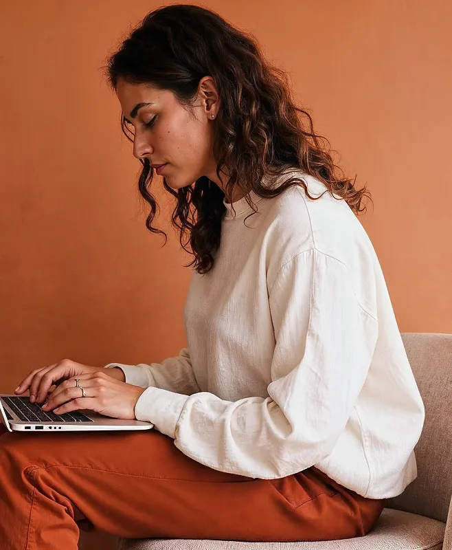 Young woman in white sweater typing on a laptop against a brown background.