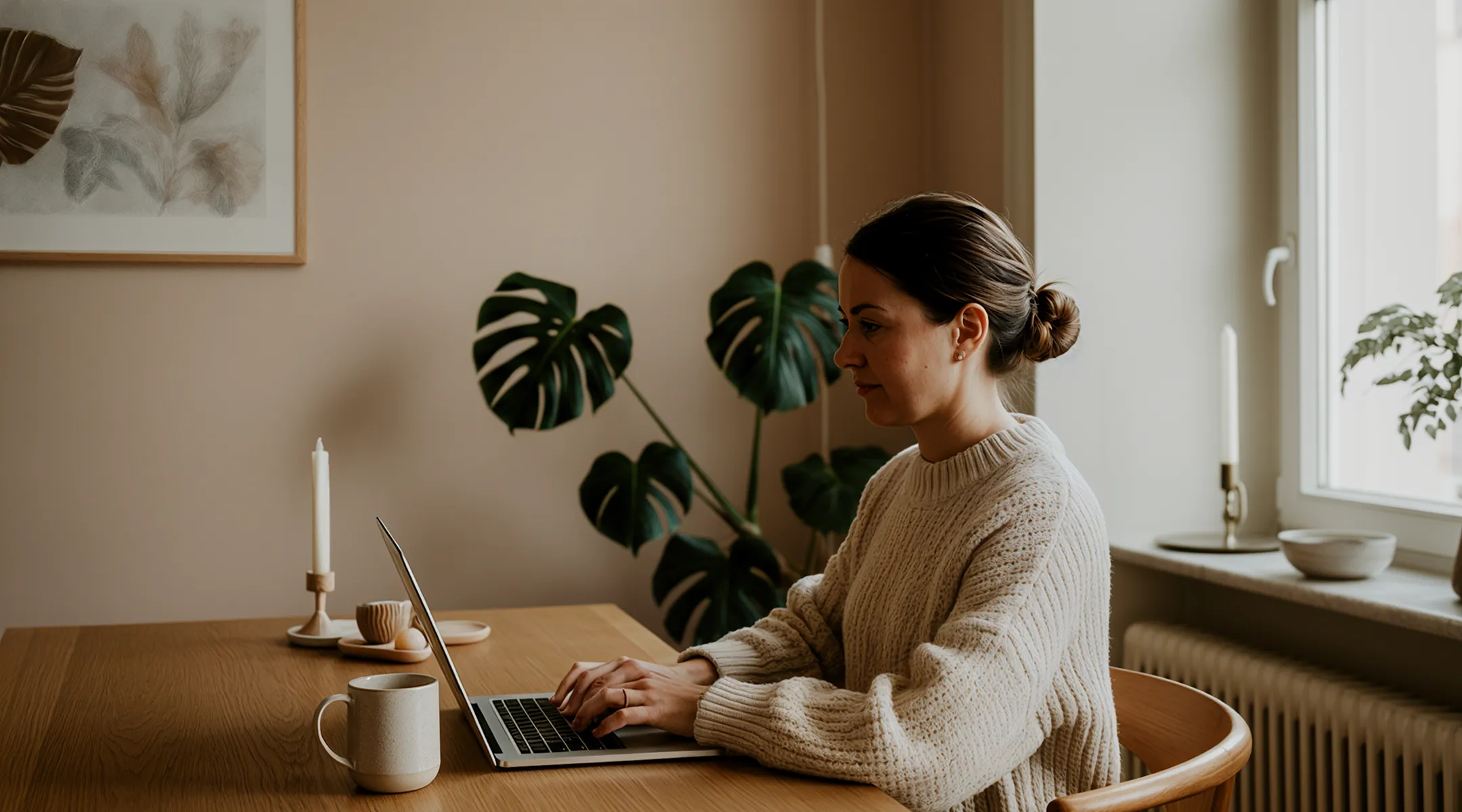 Woman in a knit sweater typing on a laptop at a wooden table with a mug and decor.
