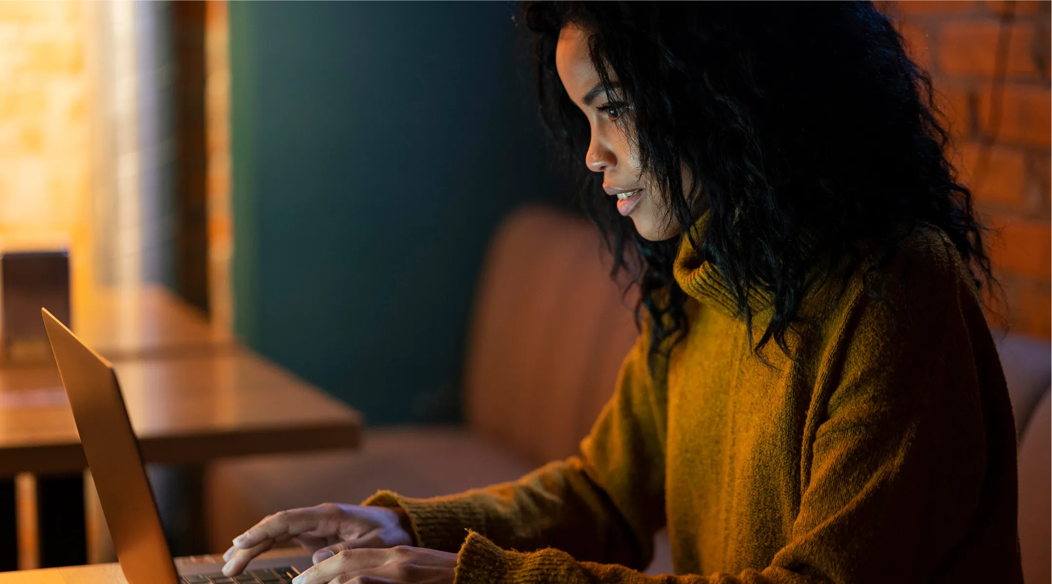 Woman in a mustard sweater focused on typing on a laptop in a cozy dimly lit room.