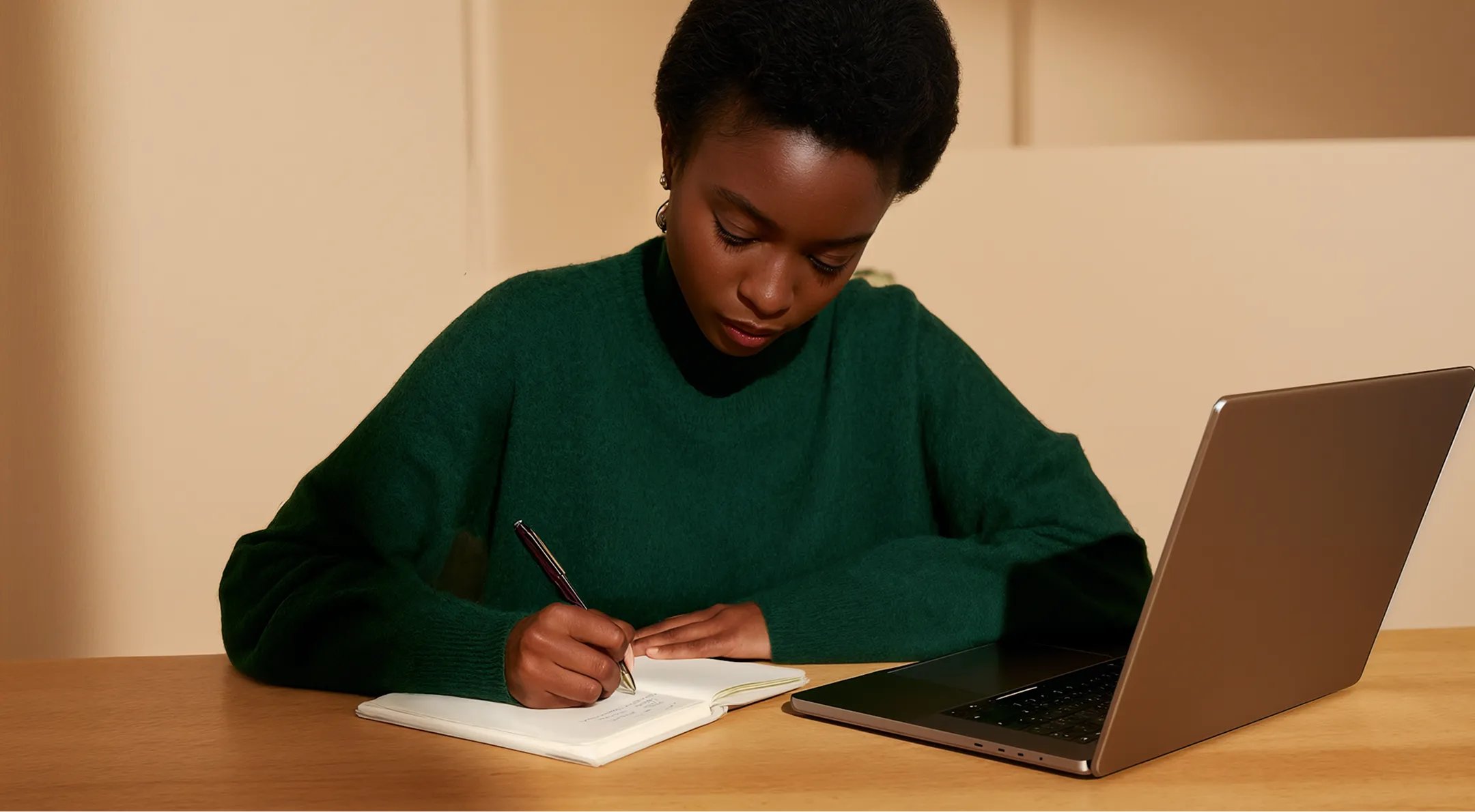 Woman in green sweater writing in a notebook beside a laptop on a wooden table.