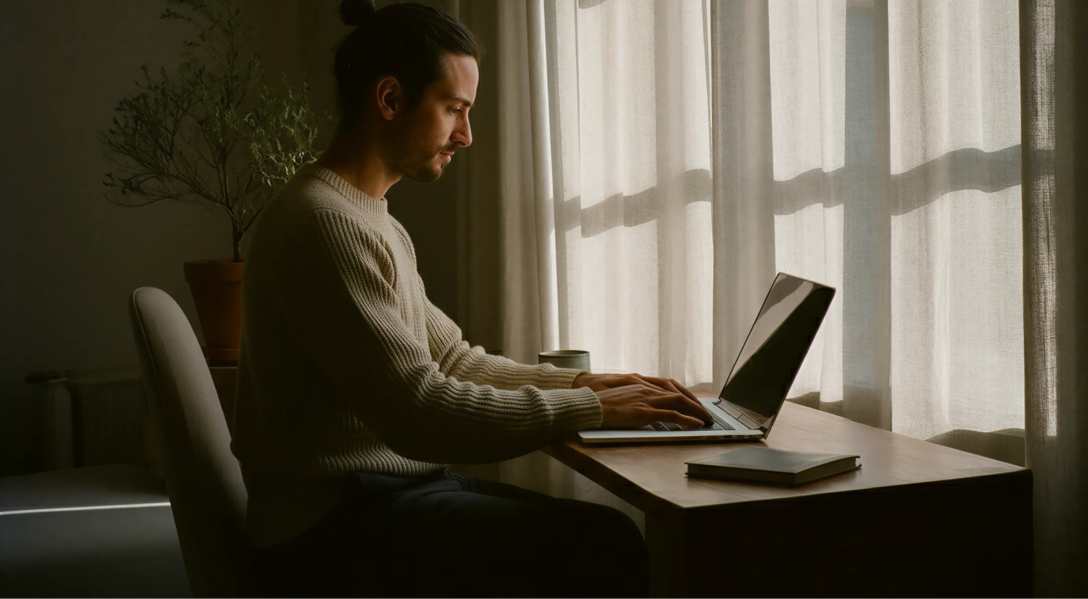 Man in a beige sweater sitting at a wooden table typing on a laptop near a window with curtains.