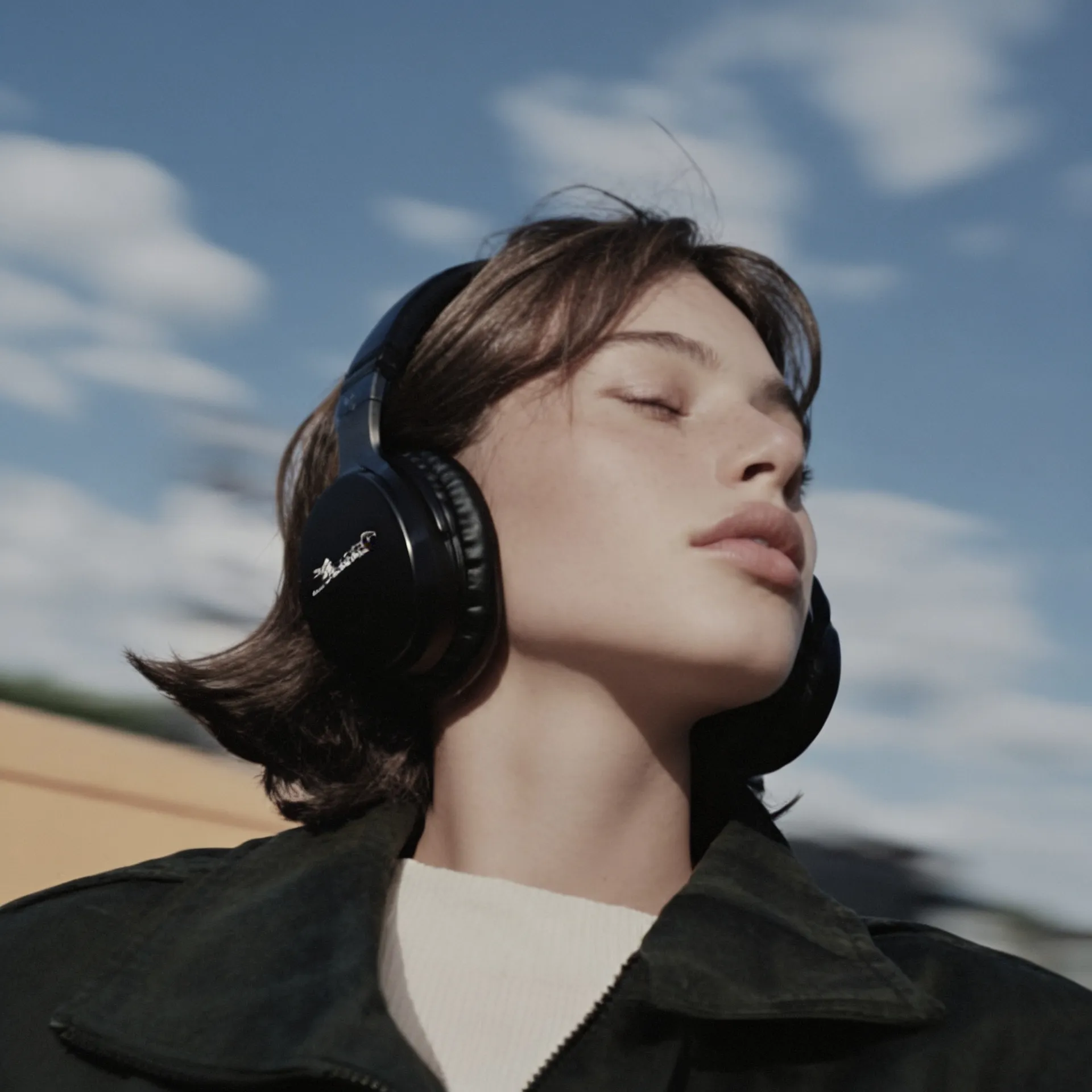 Young person outdoors wearing black headphones with eyes closed against a blue sky with clouds.